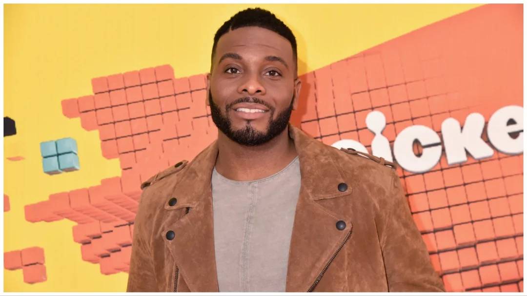 A man with short hair and a beard, wearing a brown suede jacket over a gray shirt, stands in front of an orange and yellow wall with part of the "Nickelodeon" logo visible.