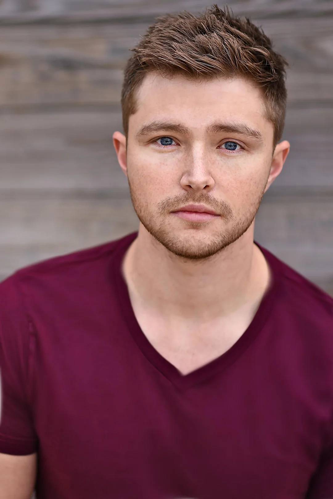 A young man with short brown hair and light facial hair wears a maroon V-neck shirt and looks directly at the camera with a neutral expression; wooden boards form the background.