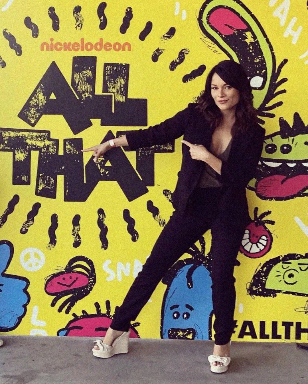 A woman in a black suit and white heels poses, pointing at a bright yellow "Nickelodeon All That" backdrop decorated with cartoon doodles and playful designs.