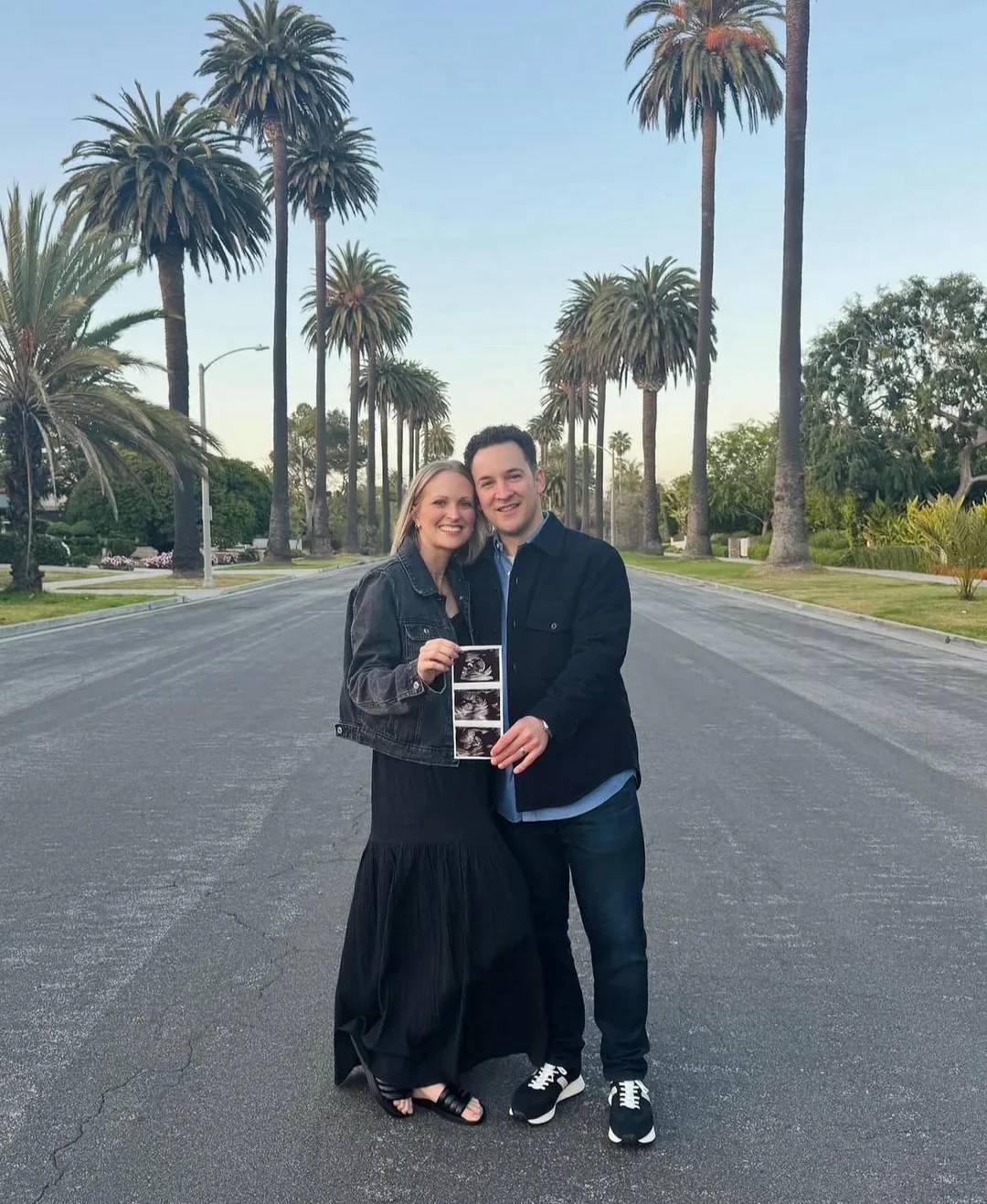 A smiling couple stands on a palm tree-lined street, holding ultrasound photos together, suggesting a pregnancy announcement. The sky is clear and both are dressed in dark clothing.