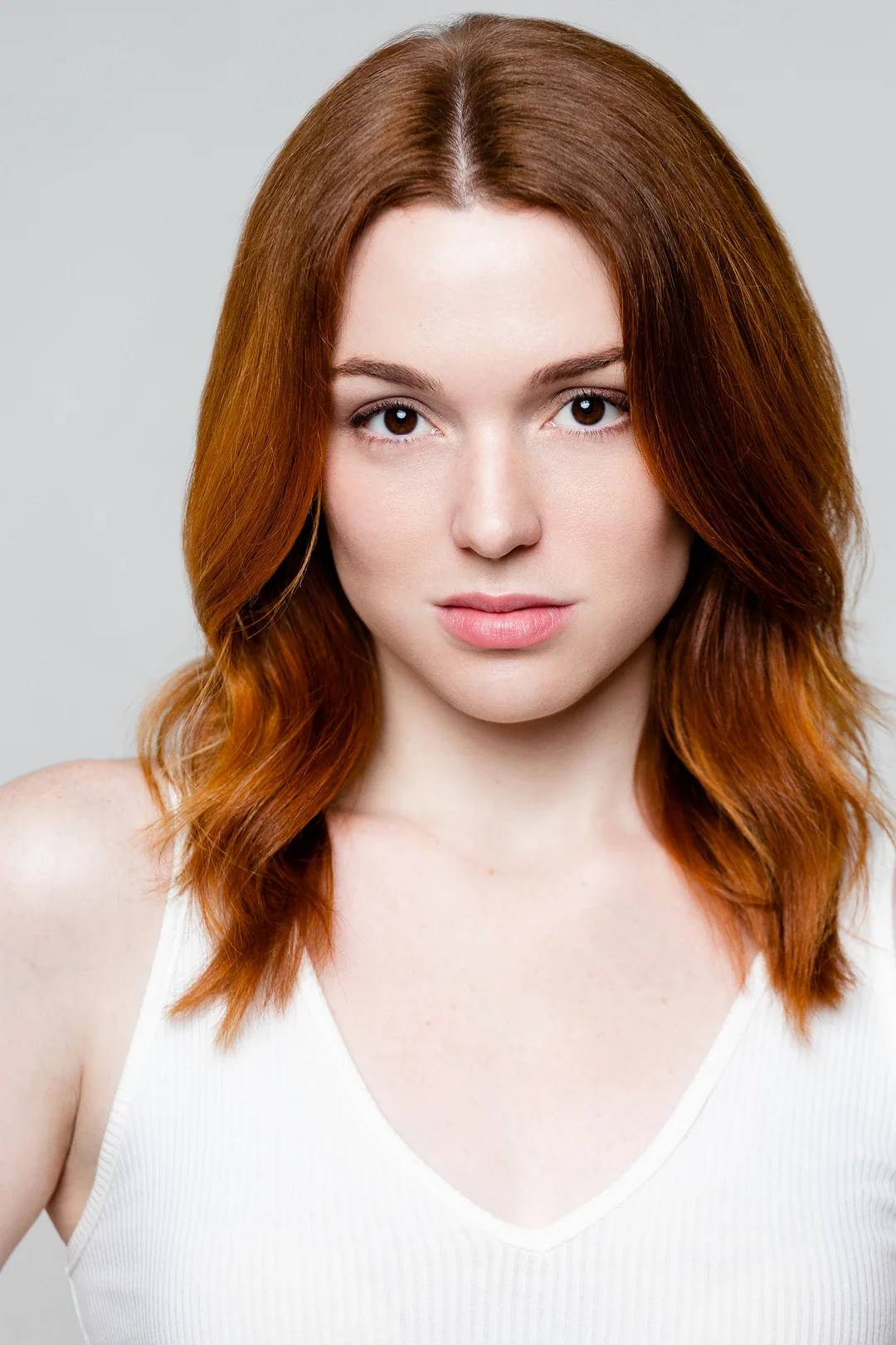 A woman with straight, shoulder-length auburn hair and fair skin looks directly at the camera with a neutral expression. She is wearing a sleeveless white top against a plain, light background.