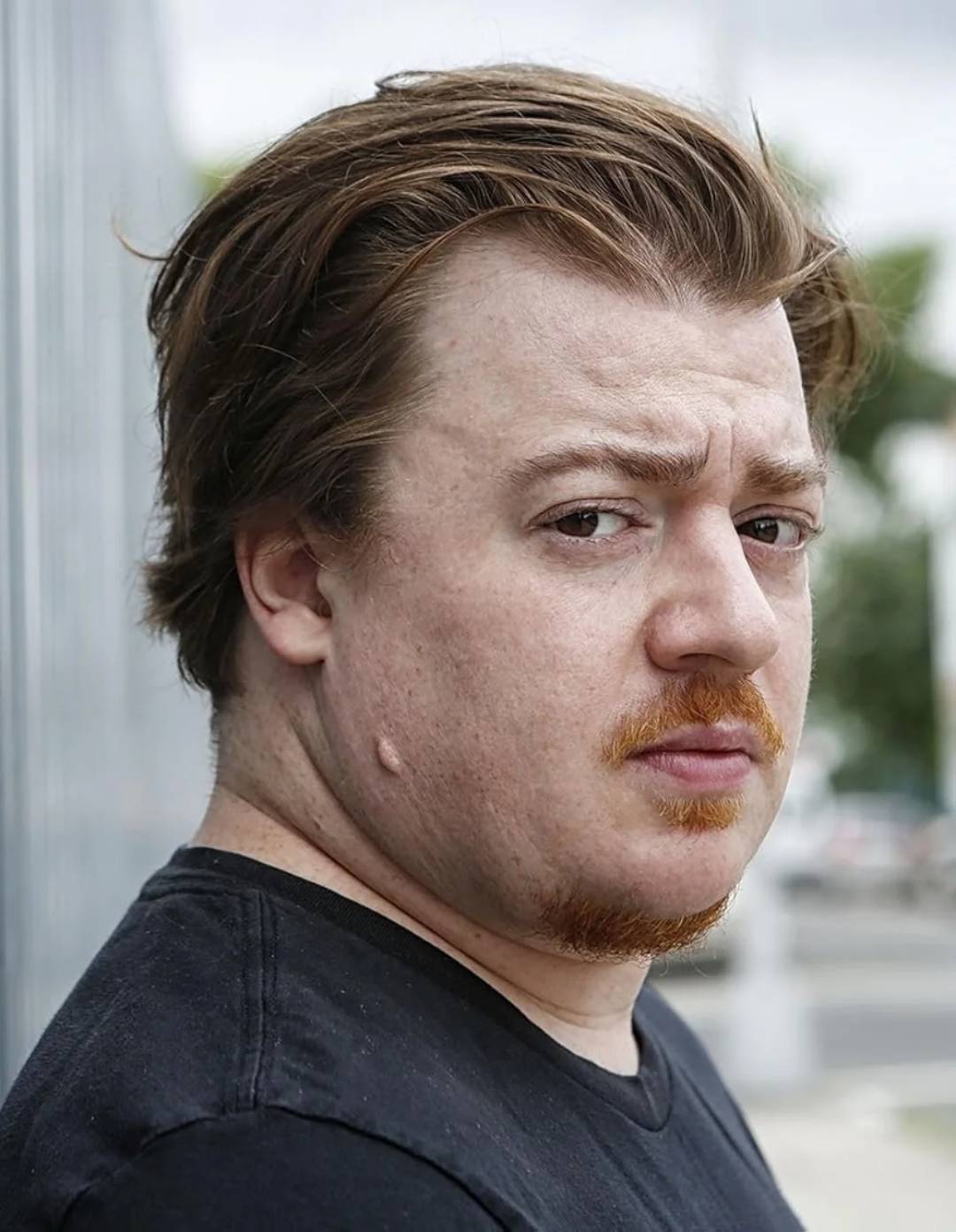 A man with light skin, reddish-brown hair, and a short reddish beard looks seriously at the camera. He is wearing a black shirt and standing outdoors near a metal fence with a blurred background.