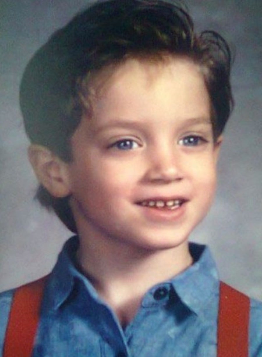 A young child with light brown hair and blue eyes smiles for a school portrait. The child is wearing a blue shirt and red suspenders, with a neutral gray background.