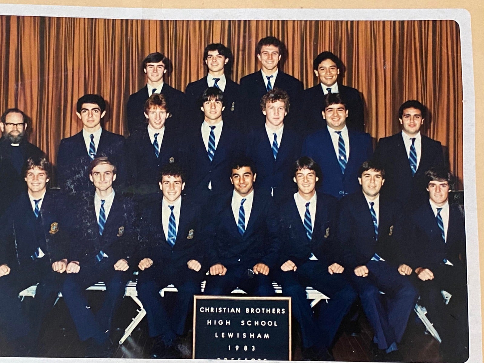 A group of young men in suits and striped ties pose for a formal class photo in front of brown curtains. A sign in front reads "Christian Brothers High School Lewisham 1983.