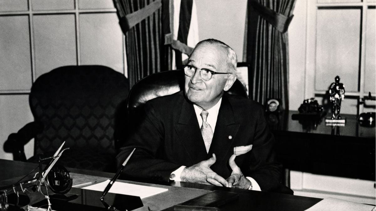 An older man in a suit and glasses sits at a large desk, smiling and looking to the side. Behind him are large curtains, flags, and shelves with various items. The image is in black and white.