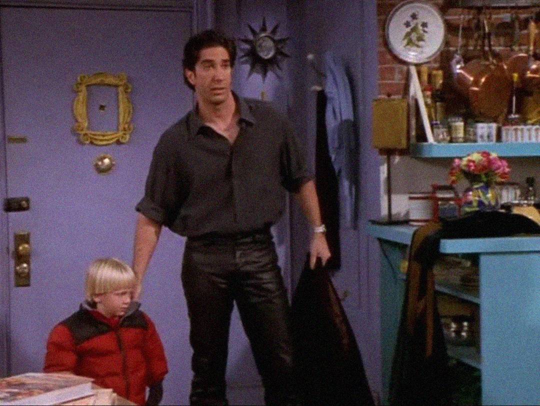A man in a dark shirt and leather pants stands holding the hand of a young blond boy in a red jacket inside a colorful, eclectic apartment with a purple door and kitchen items on shelves.