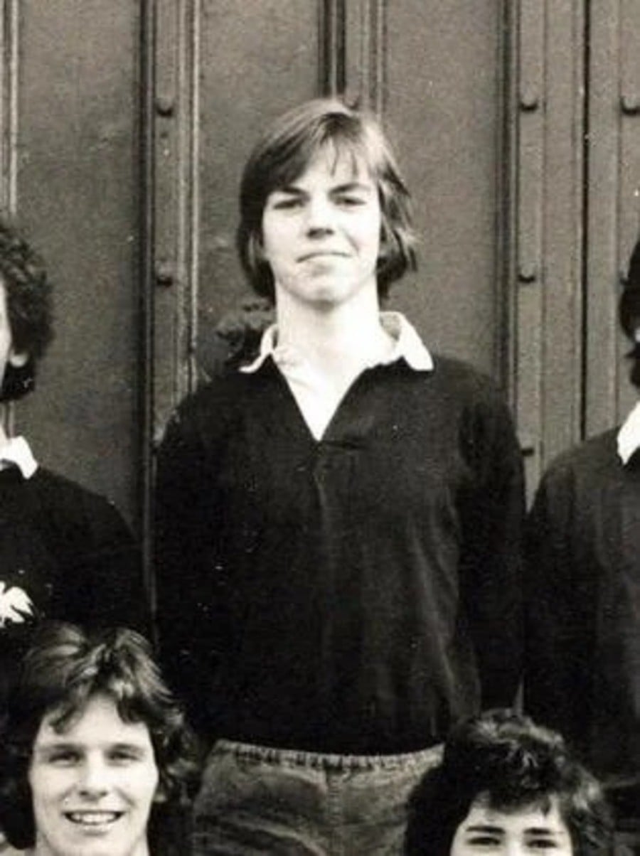 A black and white photo of a young person with medium-length hair, wearing a dark sweater over a collared shirt, standing and looking at the camera, with others partially visible around them.
