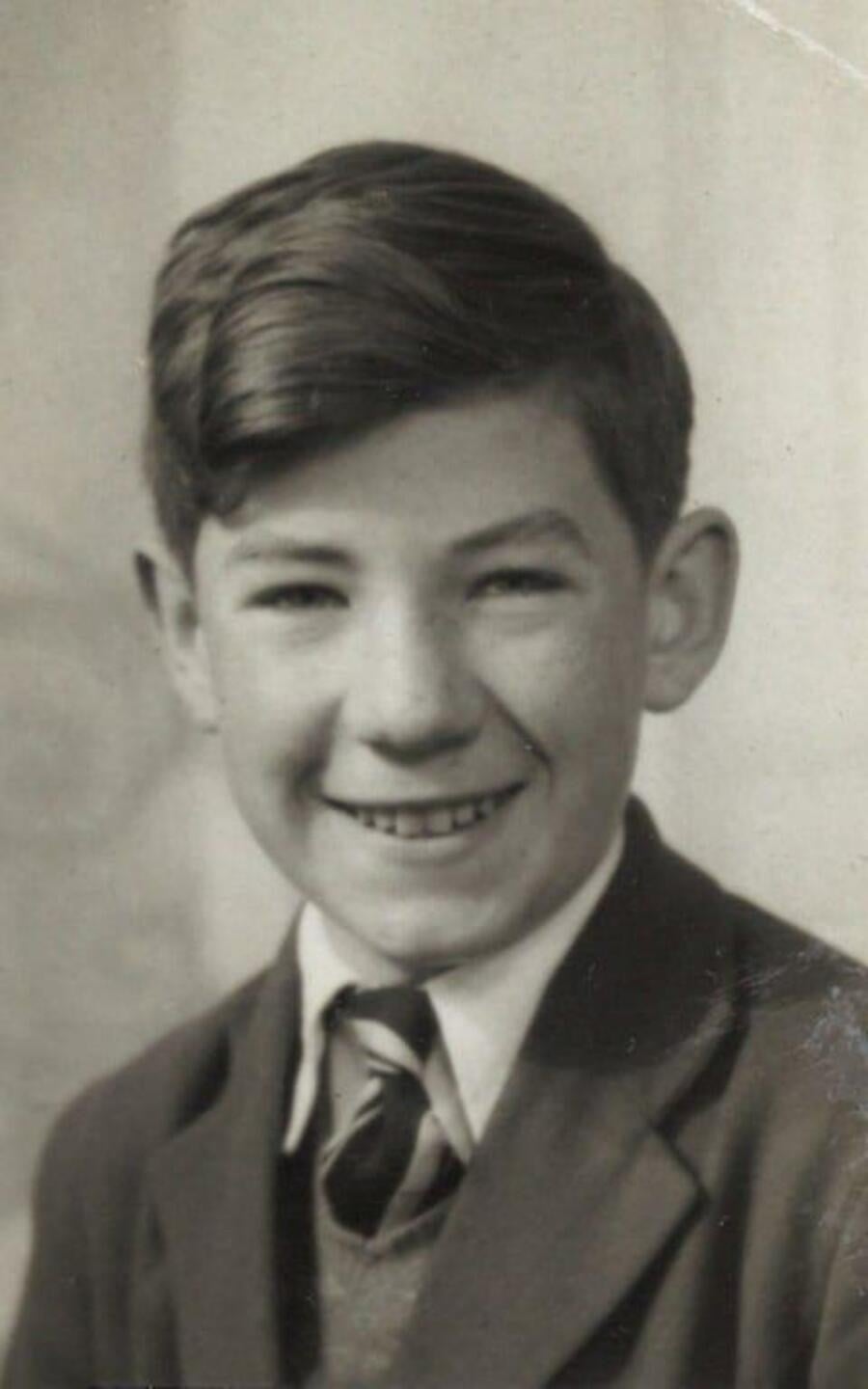 Black and white portrait of a smiling young boy with short, dark, neatly-combed hair, wearing a suit jacket, collared shirt, and striped tie. He is facing the camera against a neutral background.