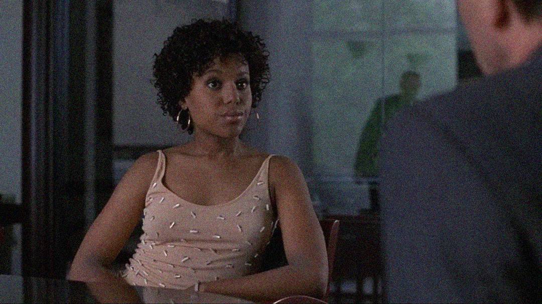 A woman with short curly hair and hoop earrings sits at a table, wearing a light-colored tank top. She faces a man whose back is to the camera, and a glass wall is in the background.