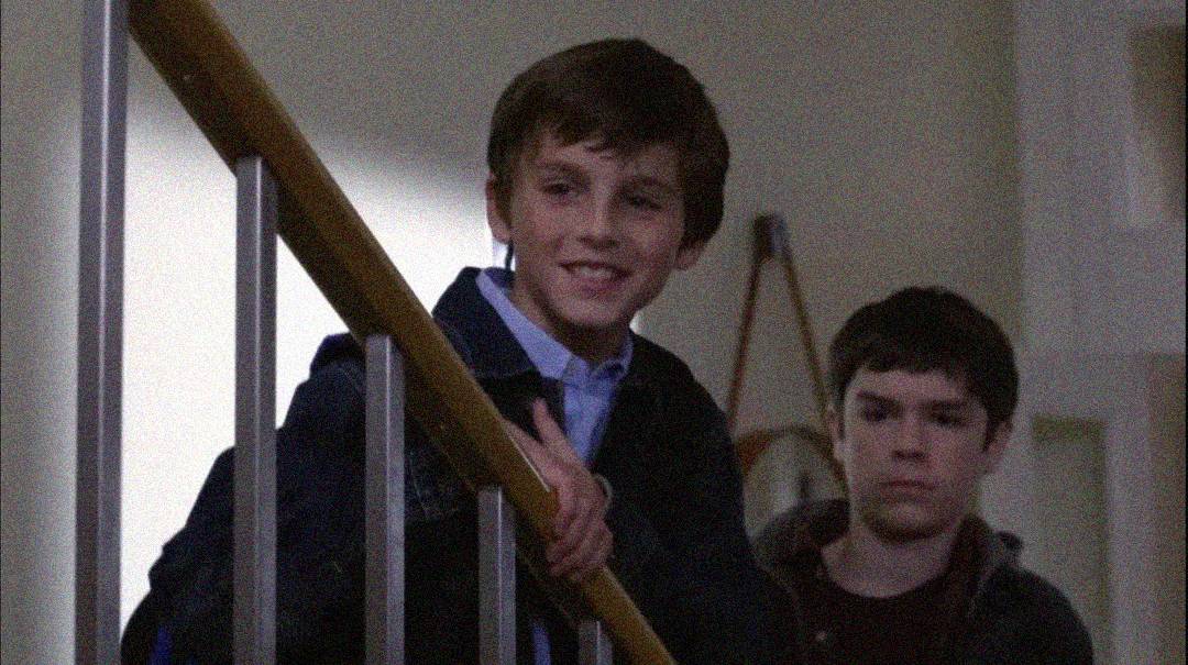 Two boys stand on a staircase indoors. The boy in front is smiling and leaning on the railing, while the boy behind him looks serious. The background appears to be a softly lit room.