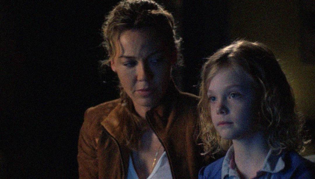 A woman with curly hair in a brown jacket sits beside a young girl with blonde hair and a blue jacket, both looking serious in a dimly lit setting.