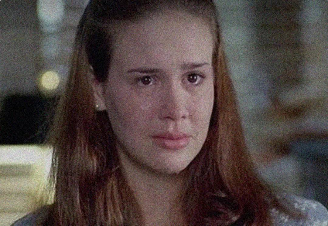 A young woman with long brown hair and teary eyes looks emotional and close to crying, staring slightly off-camera with a pained expression. The background is softly blurred.