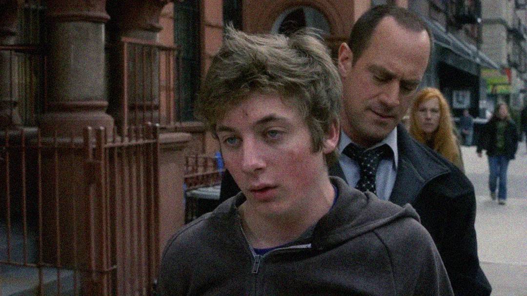 A young man with messy hair looks dazed as he is escorted by a serious-looking man in a suit on a city sidewalk, with a woman and pedestrians in the background.