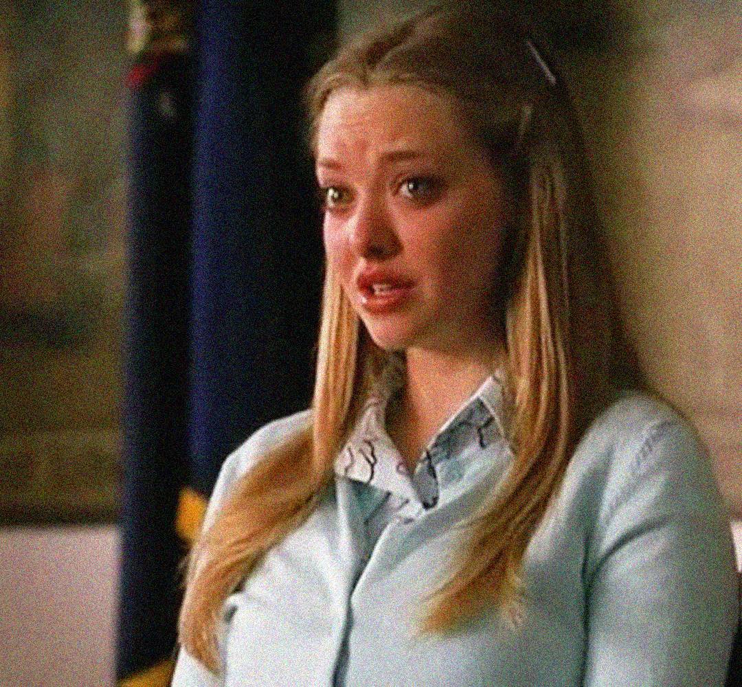 A young woman with long blonde hair, wearing a light blue cardigan and patterned blouse, stands indoors with a concerned, teary expression on her face. A dark flag is visible in the background.