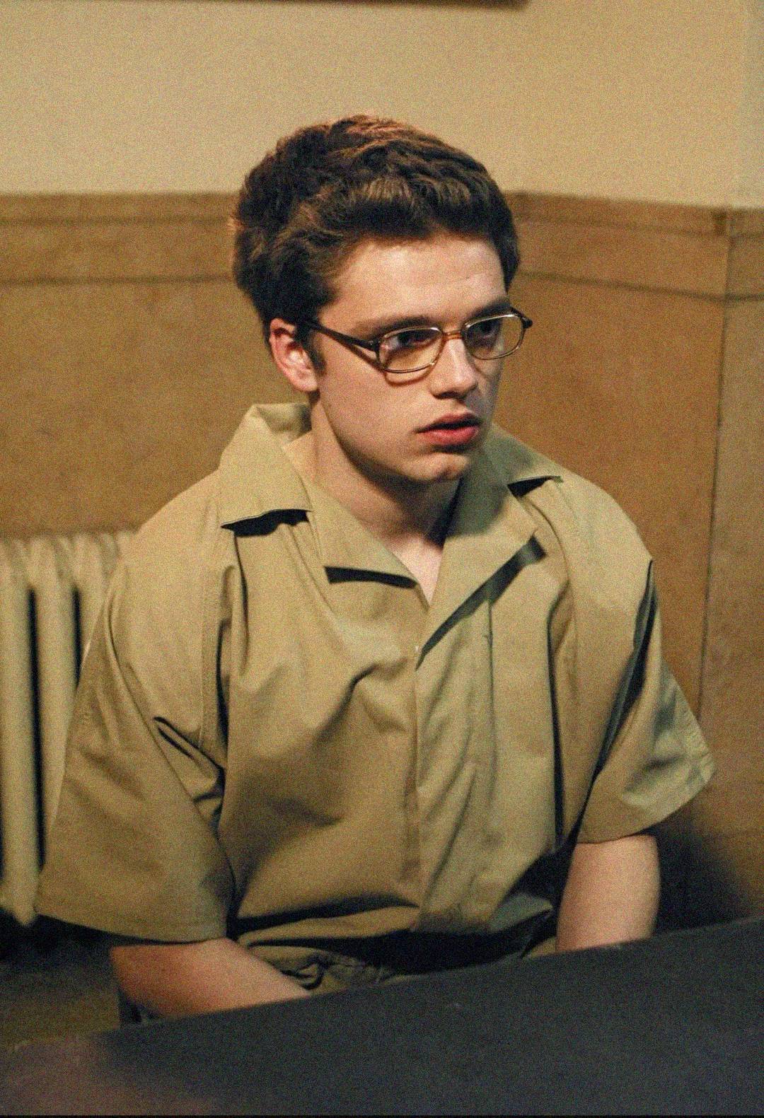A young person with short brown hair, wearing glasses and a tan prison uniform, sits at a table in a room with beige walls and a radiator in the background.