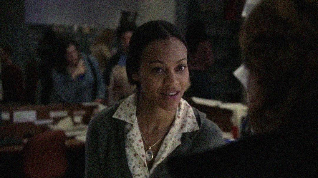 A woman wearing a white floral blouse and gray cardigan smiles while talking to someone in a dimly lit office with people and desks in the background.