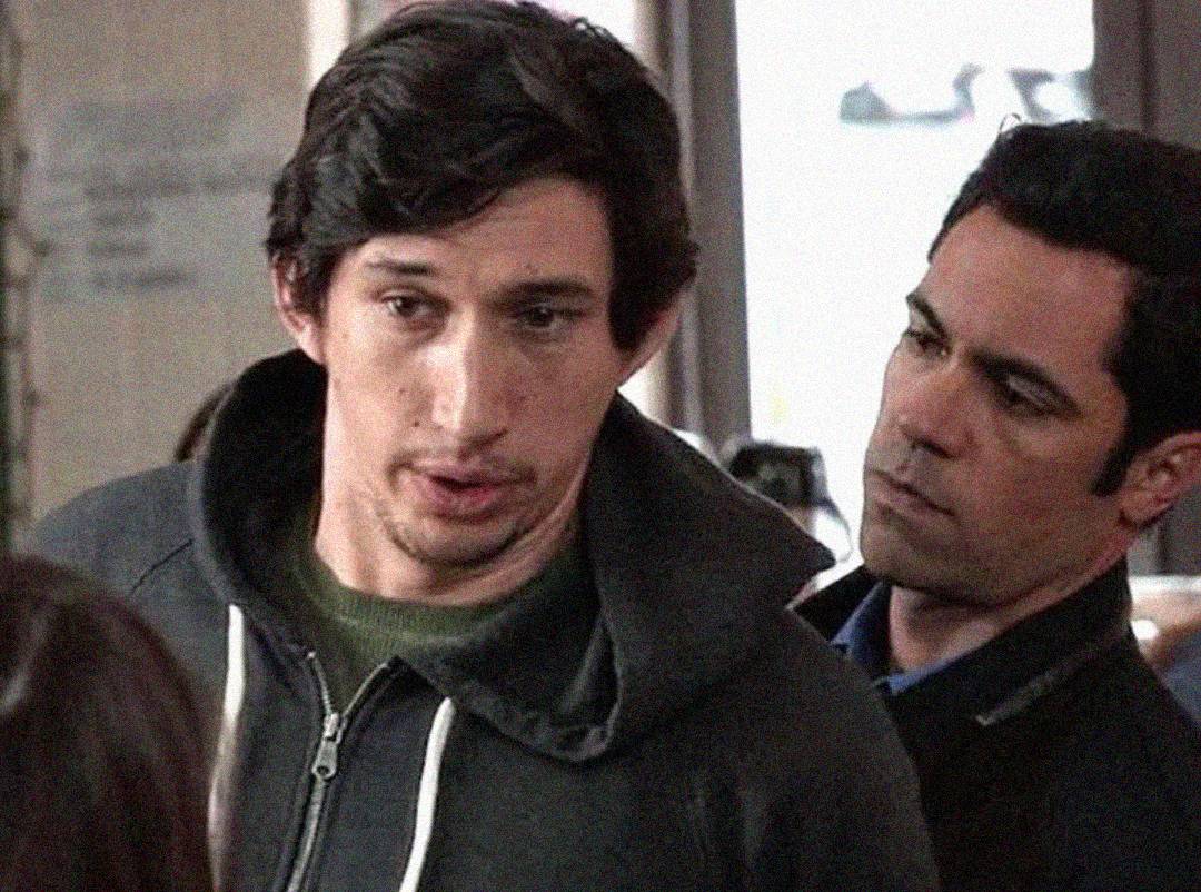 Two men stand close together indoors. The man in front wears a dark hoodie and green shirt, looking slightly to the side, while the man behind, in a black jacket, looks at him intently.