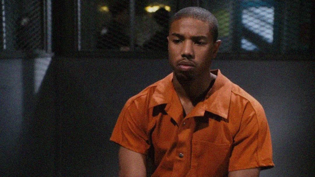 A man wearing an orange prison jumpsuit sits with a serious, somber expression in what appears to be a jail visiting area, with metal bars and a mesh screen visible behind him.