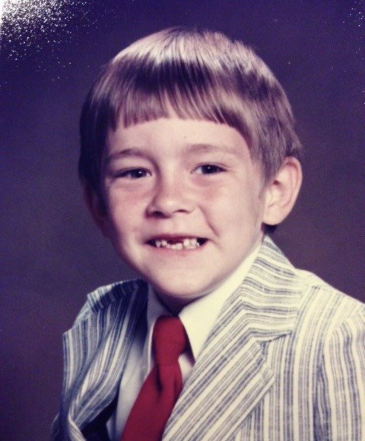 A young boy with light brown hair and missing front teeth smiles at the camera. He is wearing a striped suit jacket, white shirt, and red tie, posing against a plain, dark background.