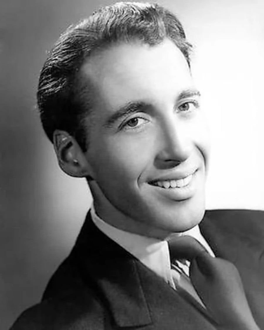 A black-and-white portrait of a young man in a suit and tie, smiling and looking slightly to the side. His hair is neatly combed, and the background is softly blurred.