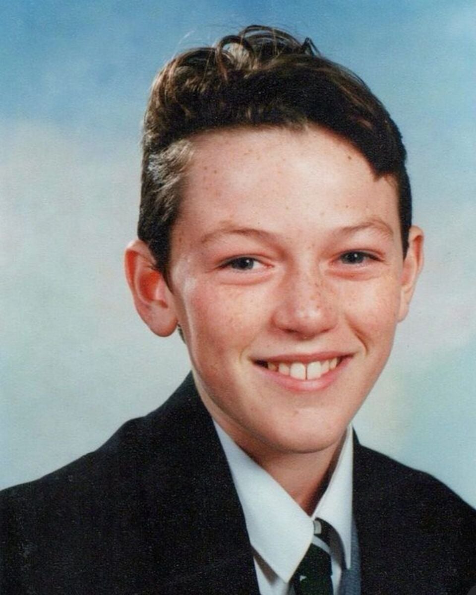 A school portrait of a young boy with fair skin, short brown hair, and freckles, smiling at the camera. He is wearing a black blazer, white shirt, and a striped tie against a light blue background.