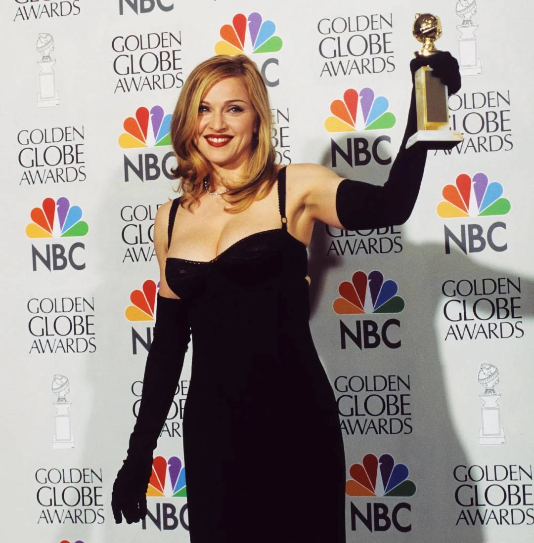 A woman in a glamorous black dress and long black gloves holds up a Golden Globe trophy, smiling, in front of a backdrop with Golden Globe Awards and NBC logos.