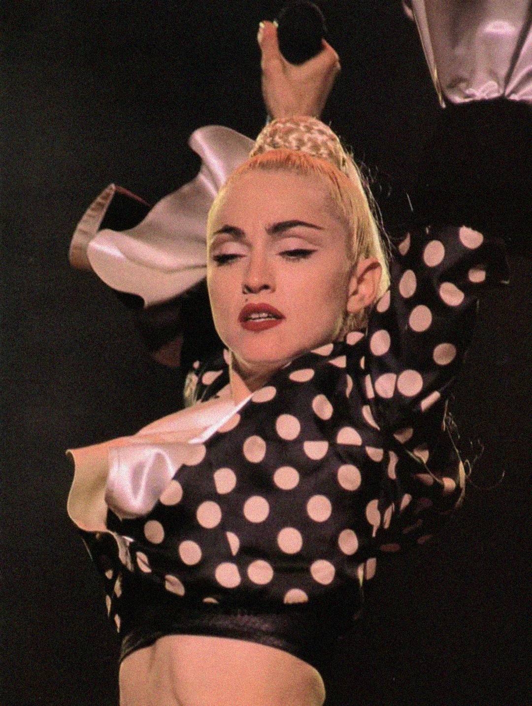 A performer with blonde hair in a high bun wears a black and white polka dot jacket. She is holding a microphone and appears to be mid-dance on stage under dramatic lighting.