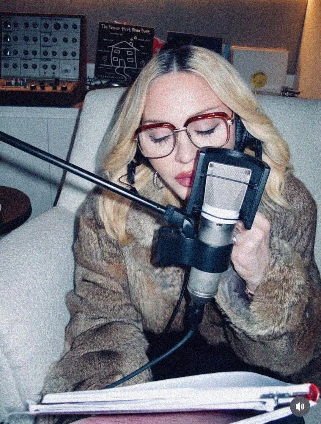 A woman with blonde hair, large glasses, and a fur coat sits in a chair, eyes closed, singing or speaking into a studio microphone with headphones on. A notebook is open in front of her. Audio equipment is visible in the background.