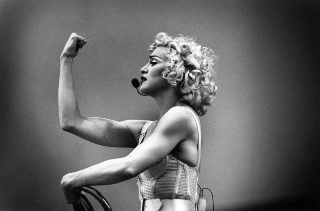 A woman with curly blonde hair, wearing a headset microphone and a cone-shaped corset, flexes her arm in a strong pose, looking upward against a plain background.