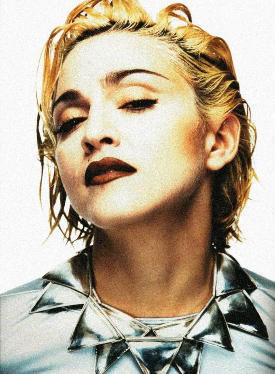 A person with short, slicked-back blonde hair and bold makeup, including dark lipstick and eyeliner, wears a metallic, geometric necklace and gazes confidently at the camera against a plain background.