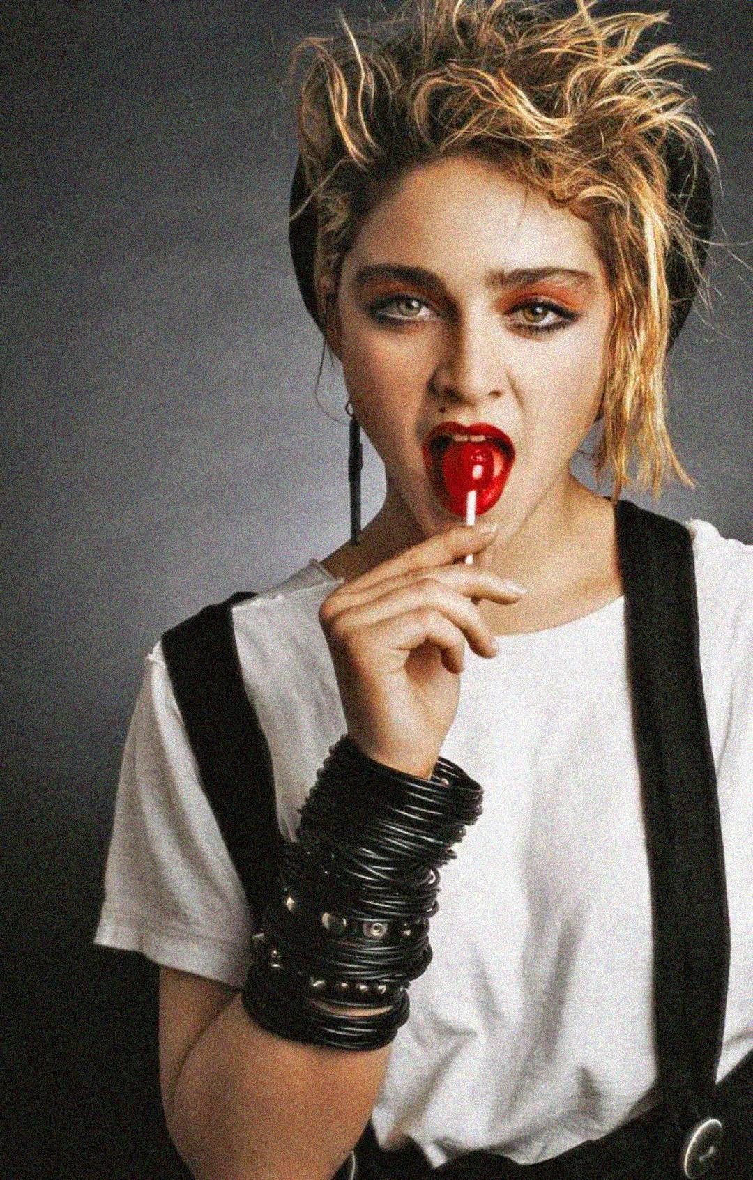 A person with messy blond hair, bold makeup, and multiple black bracelets wears a white t-shirt and black suspenders, holding a red lollipop to their mouth against a dark background.