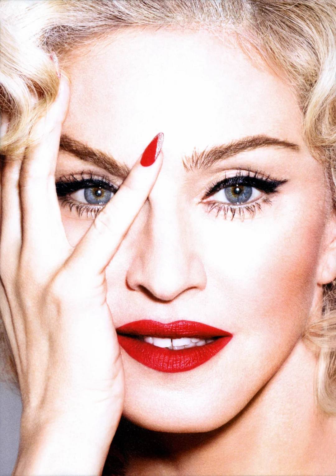 Close-up of a woman with blonde curly hair, bold red lipstick, black eyeliner, and manicured red nails. She is holding her hand to her face, with fingers partly covering one eye, looking directly at the camera.