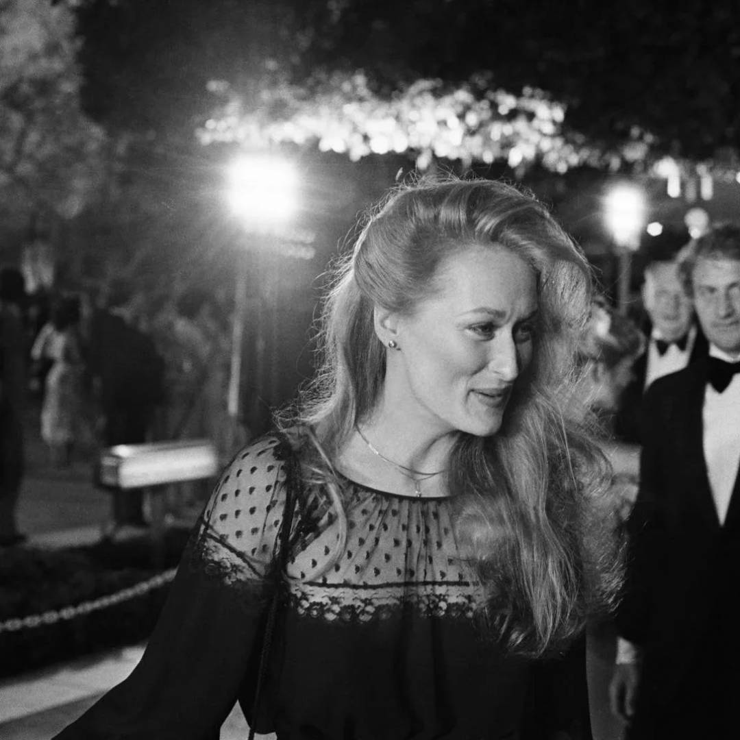A woman in a formal black dress with sheer polka-dot detailing smiles as she walks outside at night, with bright lights and people in evening wear in the background.