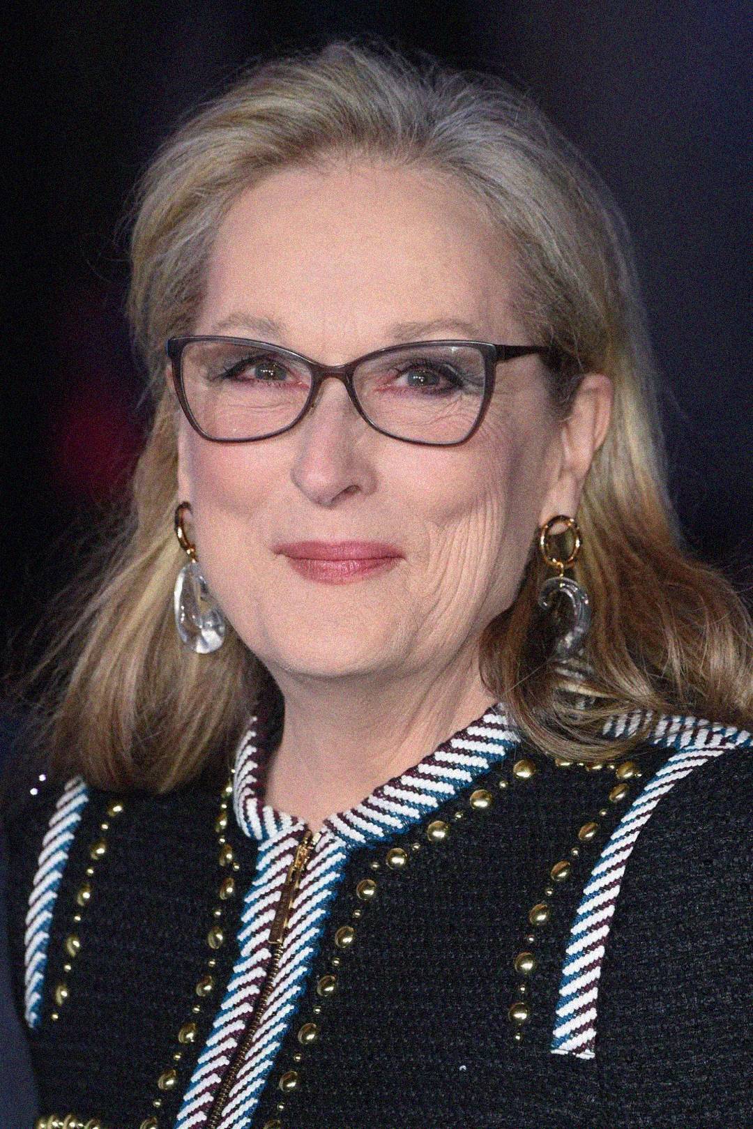 A woman with long blonde hair, wearing glasses, gold and crystal earrings, and a black jacket with studded and striped details, smiles softly at the camera.