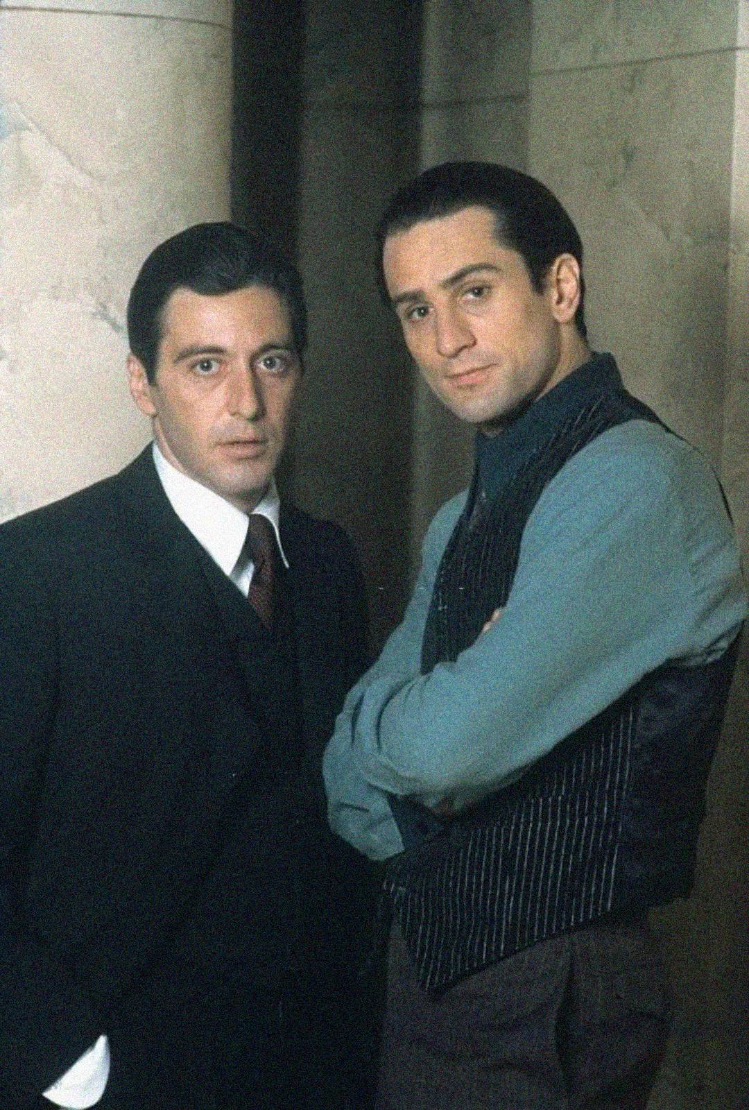 Two men in formal attire stand next to each other indoors. One wears a dark suit and tie, while the other wears a vest over a shirt with rolled-up sleeves, arms crossed, both looking at the camera in front of a marble wall.