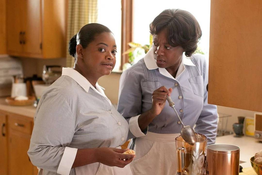Two women in 1960s-style maid uniforms stand in a kitchen. One woman holds food and looks forward, while the other stirs something in a pot and looks at her with a serious expression. Sunlight comes through the window.