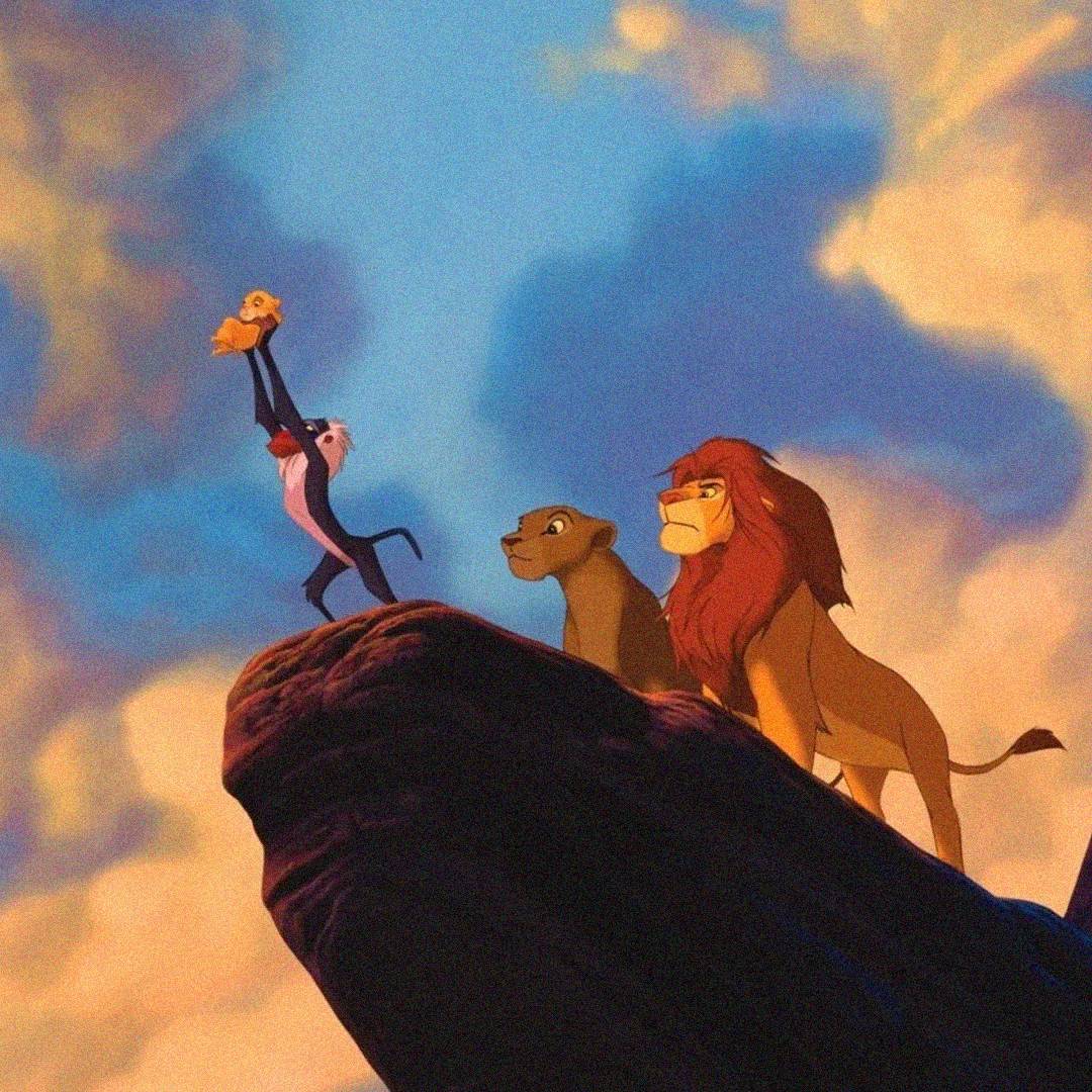 A mandrill holds a lion cub up on a rock ledge at sunrise, while two adult lions stand beside them, overlooking the savanna from "The Lion King." The sky is filled with clouds and warm sunlight.