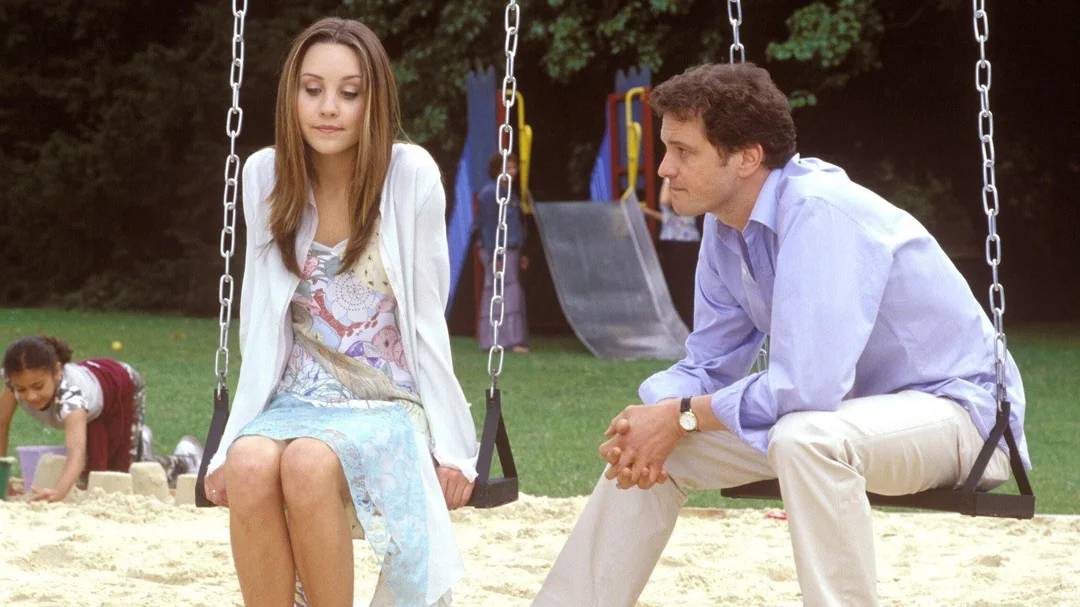 A woman and a man sit on swings in a playground, facing each other. The woman looks down with a neutral expression, while the man looks at her intently. A child plays in the sand nearby, and playground equipment is visible in the background.