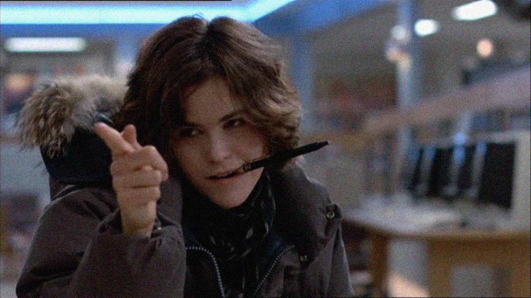 A person with short, tousled brown hair wears a dark coat with a fur-trimmed hood. They hold a pen in their mouth and gesture with their hand, finger-pointing playfully in a room with computers in the background.