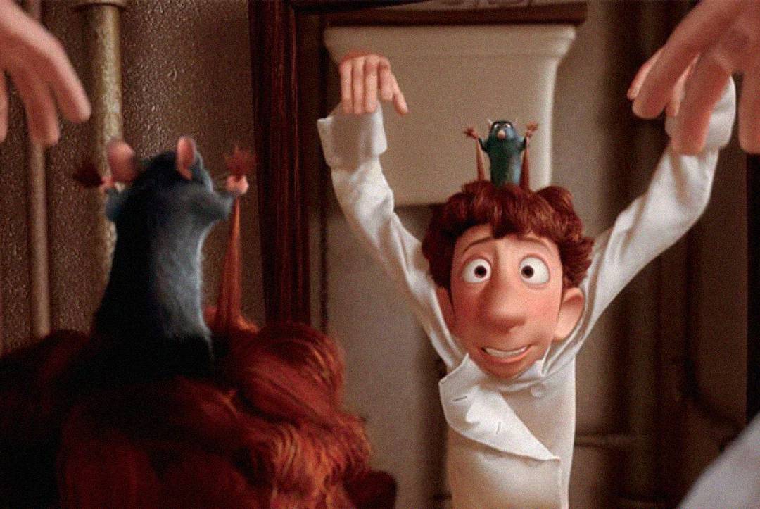 A chef in a white uniform raises his arms while a small rat sits on his head, copying his gesture. Another rat sits on a person's head in the foreground, mirroring the scene from the animated film "Ratatouille.