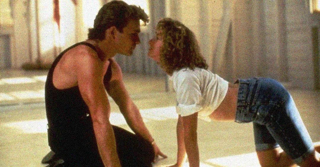 A man and woman are facing each other on the floor in a dance studio, leaning in closely as if about to kiss. The woman is on her hands and knees wearing a crop top and shorts; the man is kneeling in a black tank top.