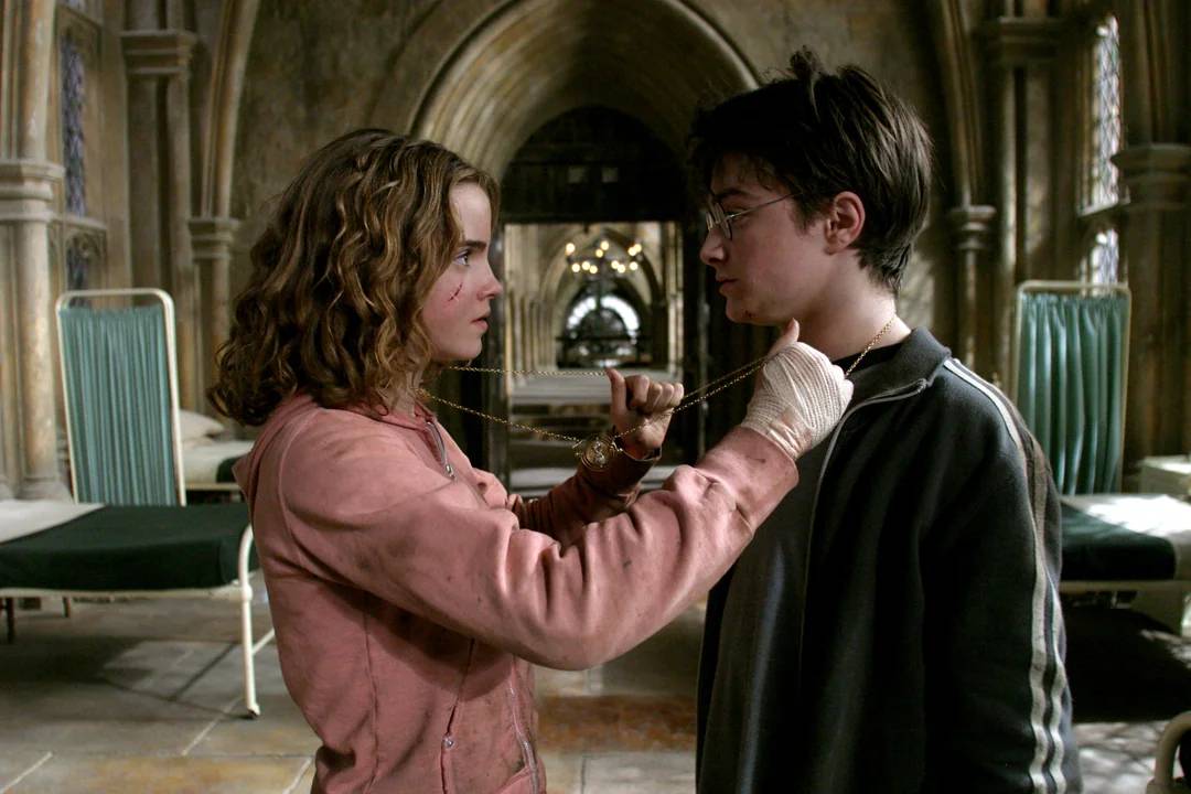 A girl with wavy hair and a pink jacket places a necklace with a time-turner around the neck of a boy with glasses and dark hair. They stand facing each other in a large, stone-walled room with empty beds.
