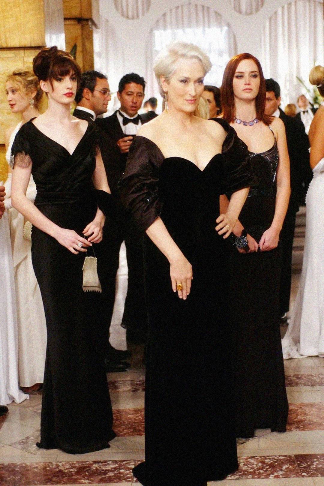 Three women in elegant black evening gowns stand at a formal event, surrounded by other guests in formal attire, in a grand, well-lit room with white drapery and marble floors.