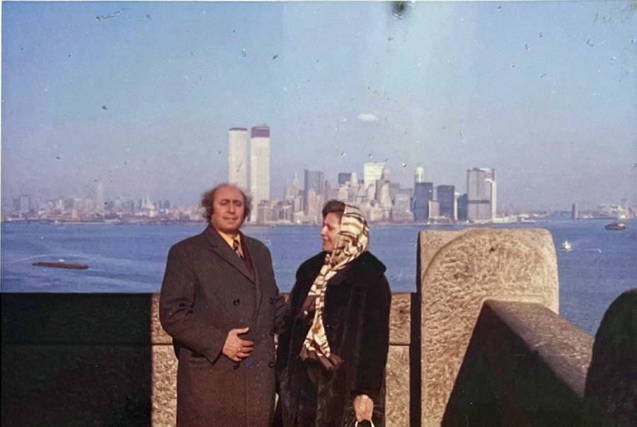 A man and a woman stand on a stone terrace overlooking the water, with the New York City skyline and the Twin Towers of the World Trade Center visible in the background on a clear day.