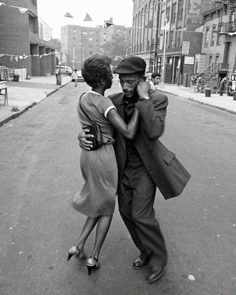 A man in a suit and hat and a woman in a dress dance closely together in the middle of an empty city street, with buildings and bunting visible in the background.