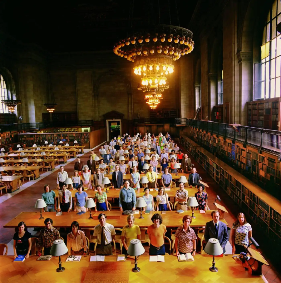A large group of people stands between rows of wooden tables in a grand library reading room with high ceilings, chandeliers, arched windows, and bookshelves lining the walls.