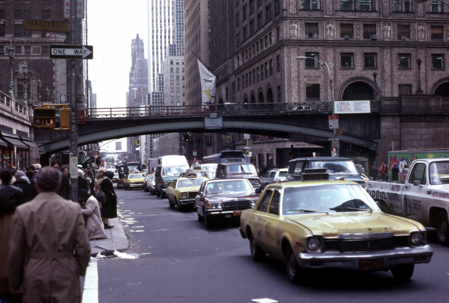 Busy city street scene with several yellow taxis, pedestrians on sidewalks, and tall buildings. An overpass bridge crosses above the traffic. The setting appears to be in New York City.