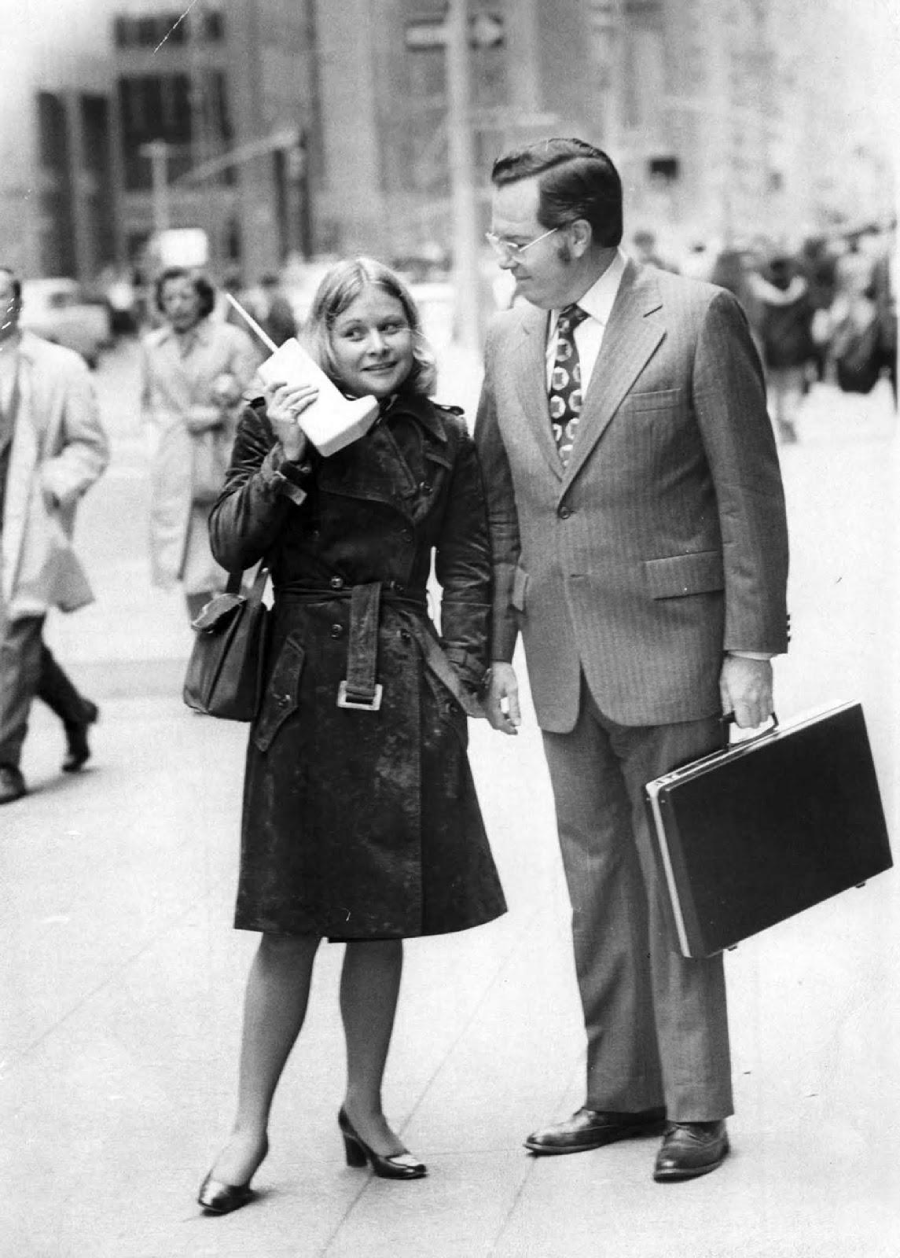 A woman in a trench coat talks on an early, large mobile phone while standing on a city sidewalk next to a man in a suit holding a briefcase. Several pedestrians walk by in the background.