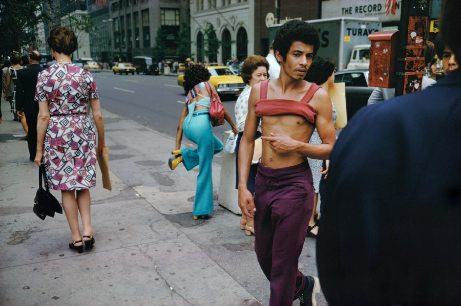 A man wearing maroon pants and a red shirt pulled up over his chest walks down a busy city sidewalk. Other people walk nearby, and cars and buildings line the street in the background.