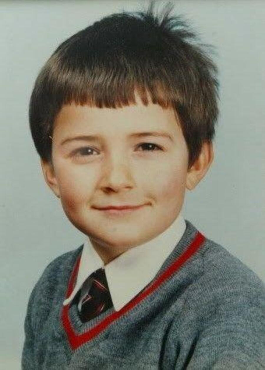 A young child with short brown hair, wearing a gray school sweater with red trim, a white shirt, and a striped tie, smiles slightly at the camera against a light background.